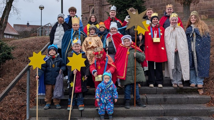 Eine Gruppe in farbenfrohen Kostümen steht auf den Kirchentreppen und hält sternförmige Requisiten während einer festlichen Veranstaltung.
