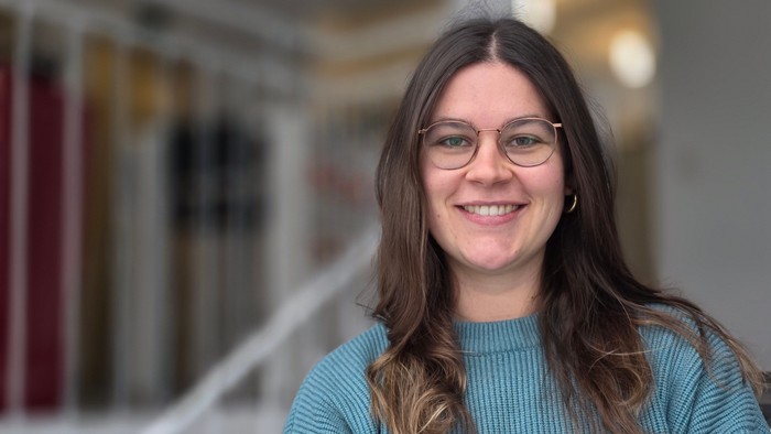 Eine Frau mit langen Haaren und runder Brille steht lächelnd vor einer Treppe, die nur unscharf zu sehen ist.