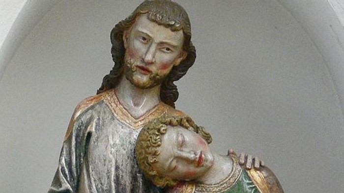 Holzstatue, die einen bärtigen Mann darstellt, der ein schlafendes Kind in traditionellen Gewändern zärtlich hält.