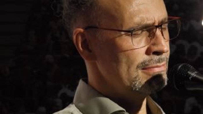 Ein Mann mit Brille und Bart, der auf einer Bühne in ein Mikrofon spricht.