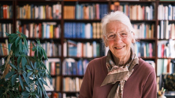 Glad ældre person står muntert i et rum fyldt med bogreoler og grønt.