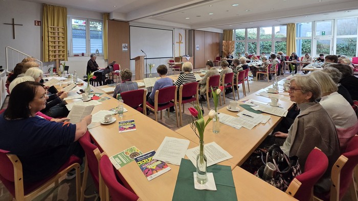 Frauen sitzen in einem Raum an Tischen und lesen in einem Heft. Vorn sitzt eine Frau am Klavier.