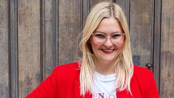 Eine fröhliche Frau in einer roten Jacke, weißem Hemd mit dem Aufdruck „VENO“ und roter Hose vor einem hölzernen Hintergrund.