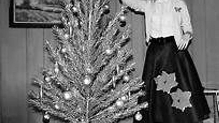 A woman in vintage attire decorates a silver Christmas tree with ornaments in a classic 1940s setting.