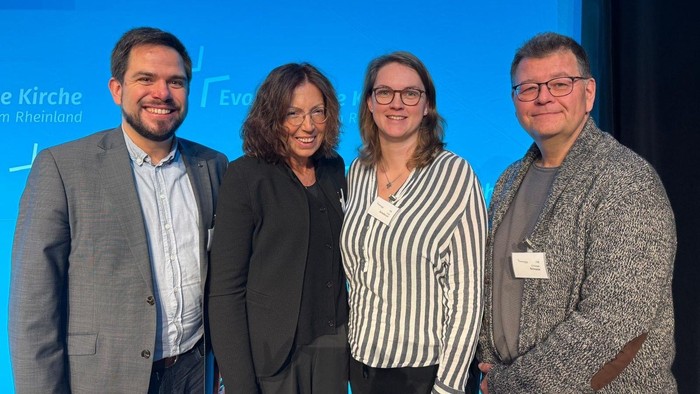 Zwei Frauen und zwei Männer lächeln in die Kamera vor einer blauen Fotowand mit Schriftzug: Ev. Kirche im Rheinland.