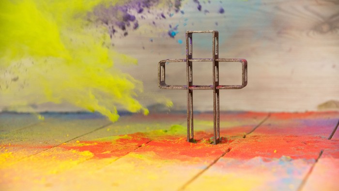 Ein kleines Metallkreuz steht auf einem bunt gefärbten Untergrund.