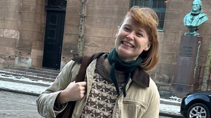 En glad person i jakke og halstørklæde, der holder en taske, står udendørs nær historisk arkitektur.