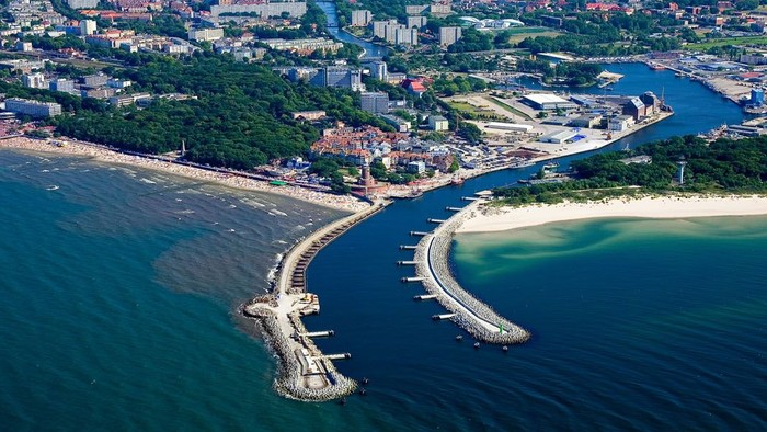 Luftaufnahme einer Küstenlandschaft mit Hafen, Strand und städtischem Panorama, bei der sich ein Wellenbrecher ins Meer erstreckt.