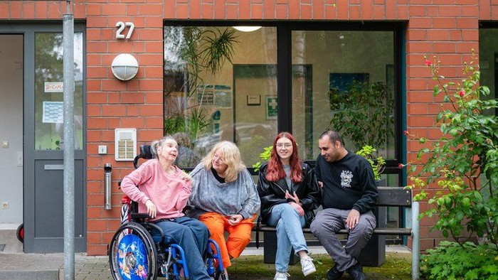 Eine Gruppe von Menschen sitzt vor einem roten Gebäude, eine Person im Rollstuhl.