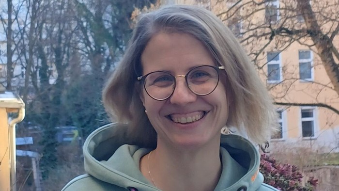 Eine lächelnde Person mit Brille, die einen grünen Hoodie trägt, steht im Freien in einer hellen, urbanen Umgebung.