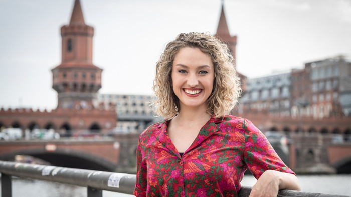 Eine fröhliche Frau in einem bunten Blumenhemd posiert vor einer Brücke mit historischen roten Backsteintürmen im Hintergrund.