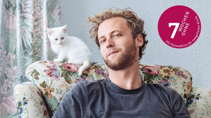 Junger Mann sitzt auf einem Plüschsessel mit Blumenmuster, auf der Kopflehne eine kleine, weiße Katze