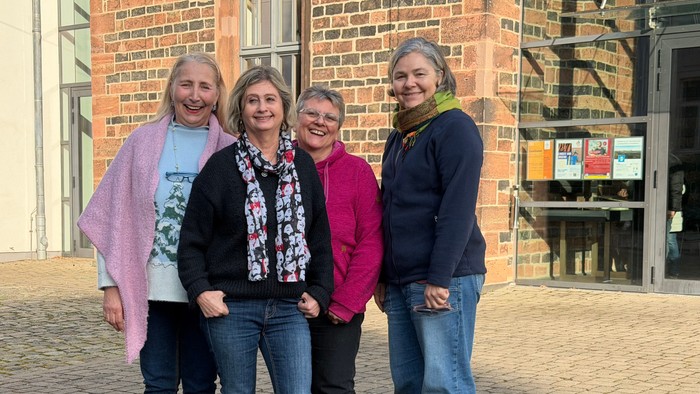 Vier Frauen, die gemeinsam draußen vor einem Backsteingebäude auf einem gepflasterten Hof lächeln.