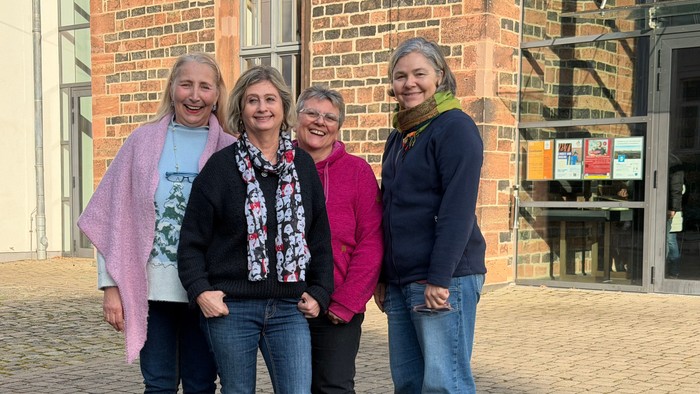 Vier Frauen, die gemeinsam draußen vor einem Backsteingebäude auf einem gepflasterten Hof lächeln.