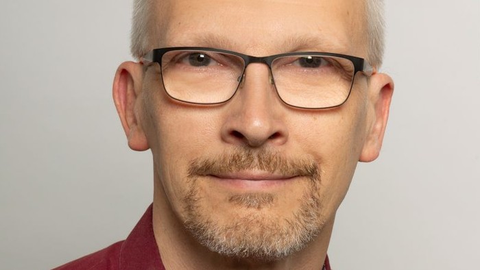 Ein Mann mit Brille und einem weinroten Hemd mit Knöpfen posiert vor einem schlichten Hintergrund.