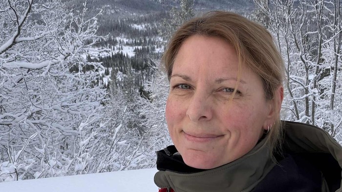 En person smiler i vintertøj foran en snedækket, skovklædt bjergbaggrund.