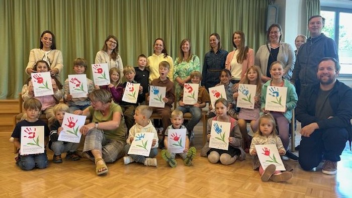 Gruppe von Kindern und Erwachsenen, die bunte bemalte Blumenbilder halten und gemeinsam drinnen posieren.