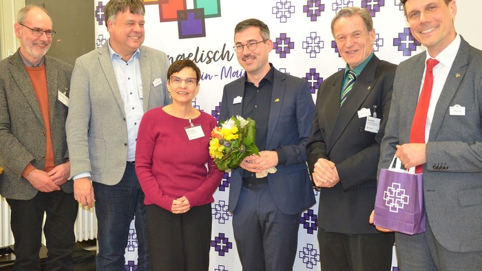 Eine Gruppe von sechs erwachsenen Personen steht nebeneinander vor einer bedruckten Fotowand mit farbigen Kreuzmotiven und dem Schriftzug „evangelisch mitten in Rhein-Main“. Die Personen sind festlich bis halbformal gekleidet, überwiegend in Sakkos und An