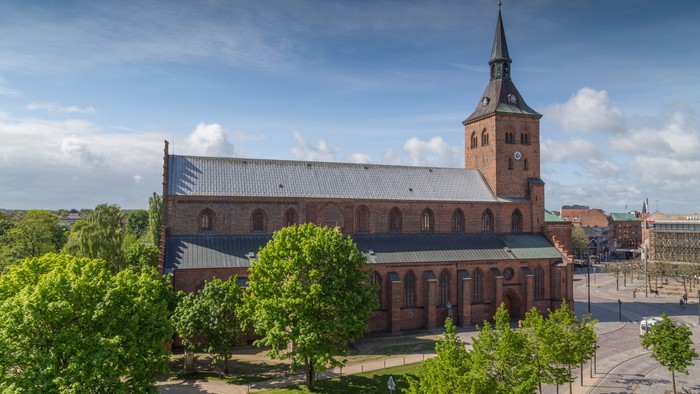 En stor kirke med spire og klokketårn, omgivet af grønne træer og et klart blå himmel.