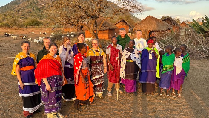Die Jugenddelegation mit jungen Massai vor einem Dorf aus Lehmbauten in einer Steppenlandschaft. Im Hintergrund ein Berg.