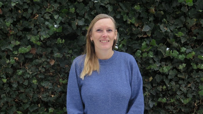En smilende person iført en blå sweater står foran en grøn hæk.