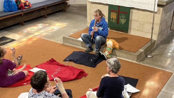 Mehrere Frauen sitzen vor einem Altar auf dem Boden einer Kirche, spielen Flöte oder machen Seifenblasen