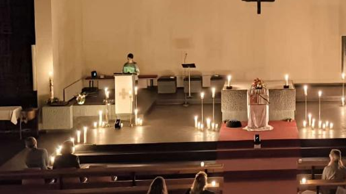 Ein ruhiger Kircheninnenraum mit brennenden Kerzen, ein Priester am Altar und Gläubige, die unten sitzen.