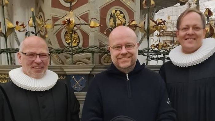 Drei Männer in religiöser Kleidung posieren gemeinsam im Inneren einer prächtigen Kirche mit aufwendigen Verzierungen.