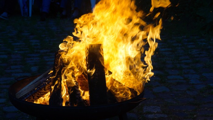 Lebhaftes Lagerfeuer, das hell in einer abendlichen Outdoor-Umgebung mit unscharfen Hintergrundfiguren brennt.