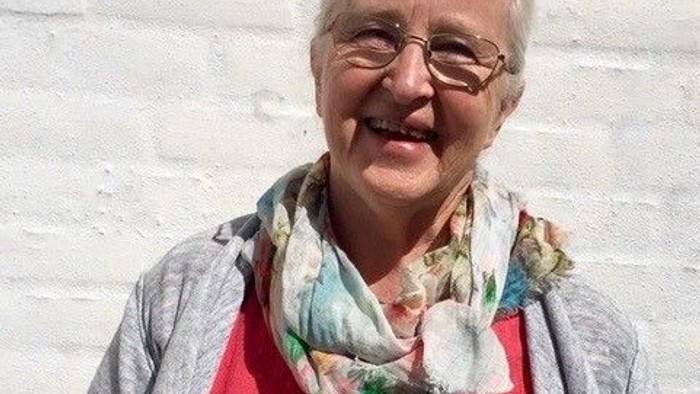 En glad kvinde med hvid hår og briller står foran en hvid mur.