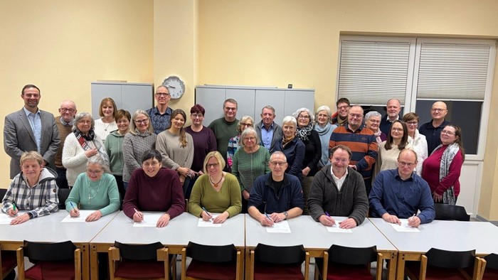 Gruppe von Personen, die gemeinsam in einem Klassenzimmer oder Besprechungsraum mit Papieren auf den Tischen posieren.