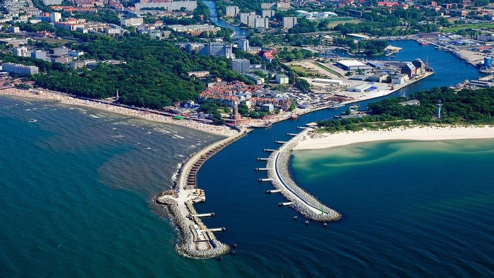Luftaufnahme einer Küstenlandschaft mit Hafen, Strand und städtischem Panorama, bei der sich ein Wellenbrecher ins Meer erstreckt.