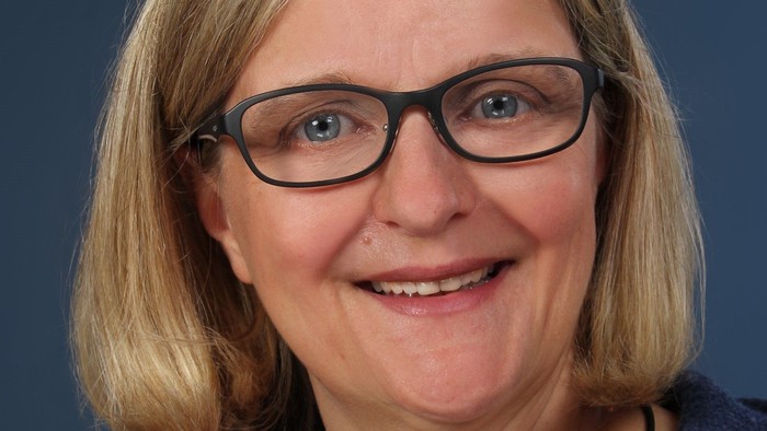 Professionelle Frau mit Brille, lächelnd, in einem dunkelblauen Cardigan über einem weißen Oberteil.