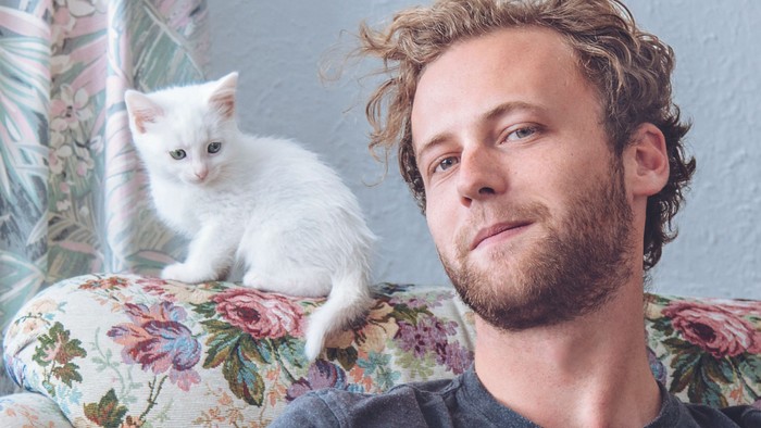 Junger Mann sitzt auf einem Sessel, neben ihm sitzt eine weiße Katze.