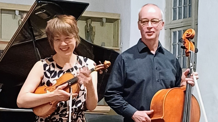 Zwei Musiker, eine Geigerin und ein Cellist, posieren gemeinsam neben einem Flügel in einem Konzertsaal.