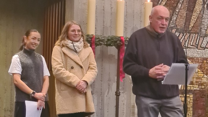 Drei Personen stehen in einer Kapelle, wobei eine eine Tafel hält und spricht, während die beiden anderen neben Kerzen zuhören.