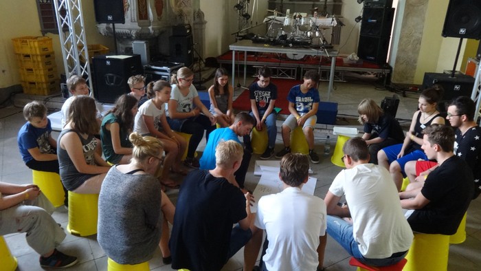 Eine Gruppe von Menschen, die während einer interaktiven Indoor-Sitzung oder eines Workshops im Kreis auf gelben Hockern sitzt.