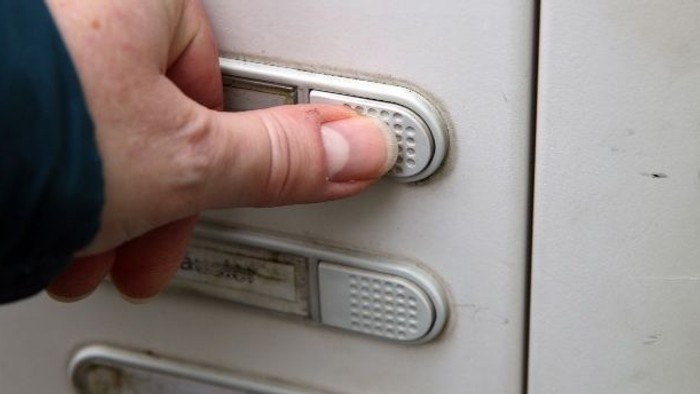 Eine Hand, die auf einem weißen Türsprechanlagen-Panel mit zwei beschrifteten Tasten Knöpfe drückt.