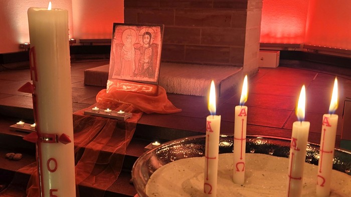 Ein mit Kerzenlicht erleuchteter Altar mit fünf brennenden Kerzen in einer Metallschale, gerahmt von religiöser Kunst und einer hohen nummerierten Kerze.