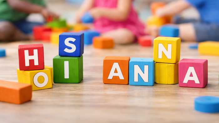 Bunte Holzklötze, die „H O I S A N A“ buchstabieren, mit spielenden Kindern im Hintergrund.