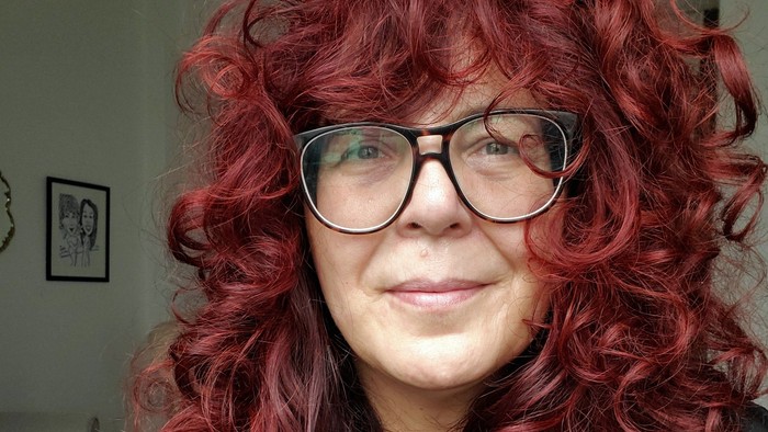 Person mit üppigem roten Lockenhaar und Brille, die eine dunkle Jacke trägt.