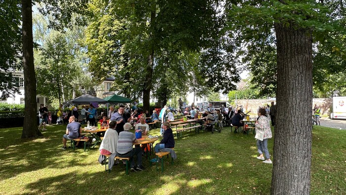 Draußen versammelte Menschen, die an Picknicktischen unter schattigen Bäumen in einer Parkanlage sitzen.
