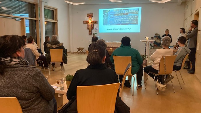 Eine Gruppe von Menschen versammelte sich drinnen und verfolgte aufmerksam eine Präsentation oder Diashow auf einem Bildschirm in einer kirchenähnlichen Umgebung.