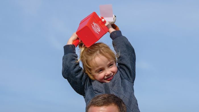 En glad mand bærer et barn, mens han holder en rød beholder, og promoverer en fællesskabsindsamling den 8. marts.