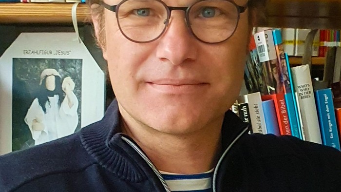 Ein Mann mit Brille und einem hohen blonden Scheitel vor einem Bücherregal