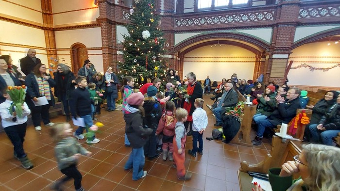 In einer Kirche versammeln sich nach dem Gottesdienst sich viele Kinder und Erwachsene um eine Frau, die Blumen entgegennimmt, während die Gemeinde applaudiert und eine warme Abschiedsstimmung herrscht.

