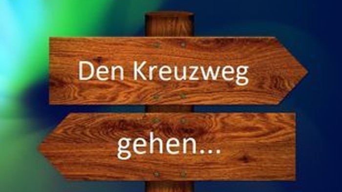 Holzwegweiser mit deutschem Text *„Den Kreuzweg gehen…“* vor unscharfem Hintergrund.