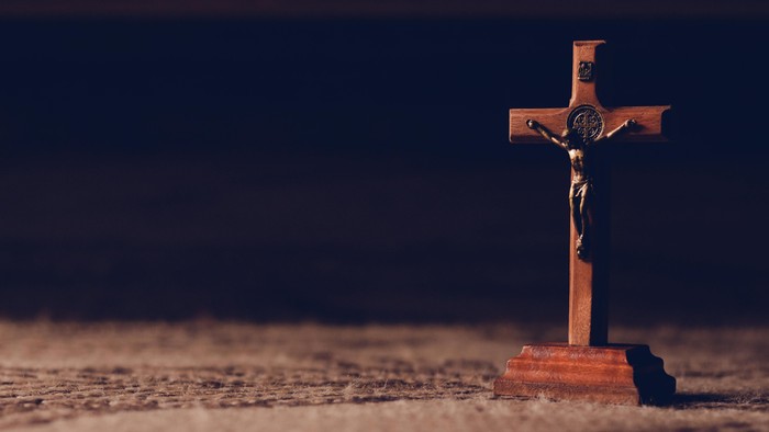 Ein hölzernes Kruzifix mit einer geschnitzten Figur steht auf einer strukturierten Oberfläche vor dunklem Hintergrund.