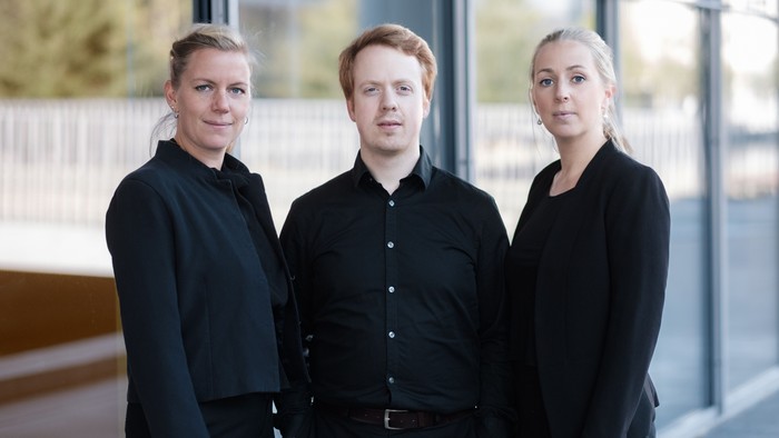 Tre fagfolk i formelt sort tøj, der poser sammen indendørs nær store vinduer.