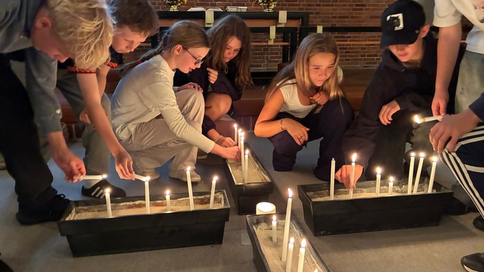 En gruppe mennesker tænder lys i lange lysestager under en indendørs sammenkomst eller ceremoni.